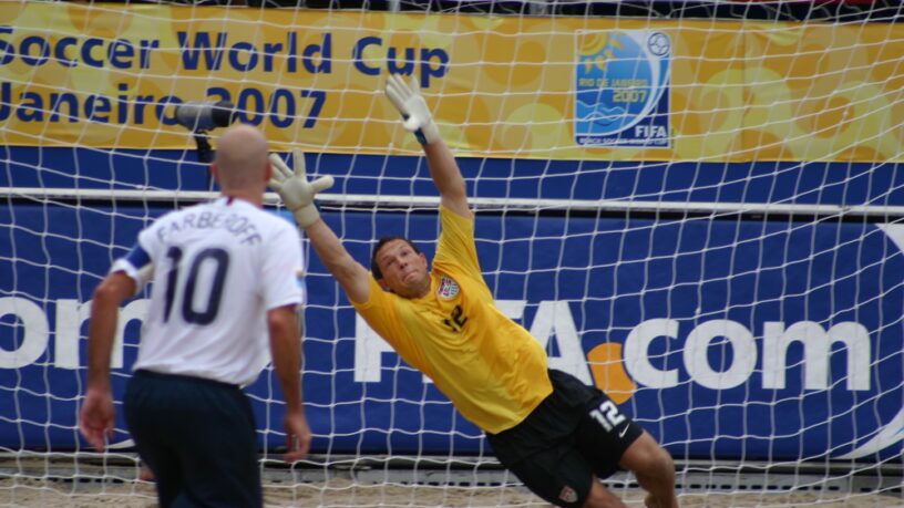beach soccer goalie jumps to catch ball before it hits the goal